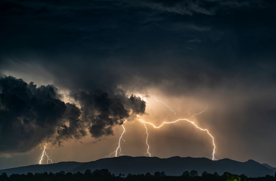 Spectacular lightning strikes illuminate dark clouds over a mountain range during a thunderstorm.