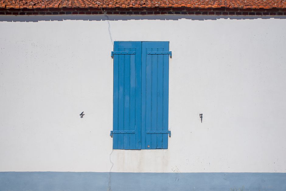Simple minimalistic exterior with a closed blue shutter on a white wall and red roof.
